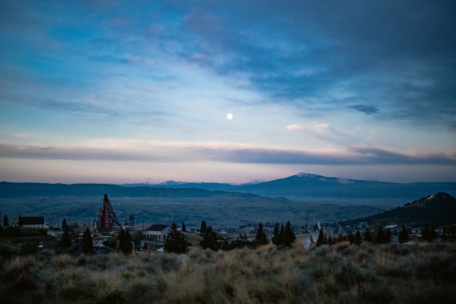 The Butte valley at dusk beneath a rising moon, a historic headframe standing above the town with the mountains beyond