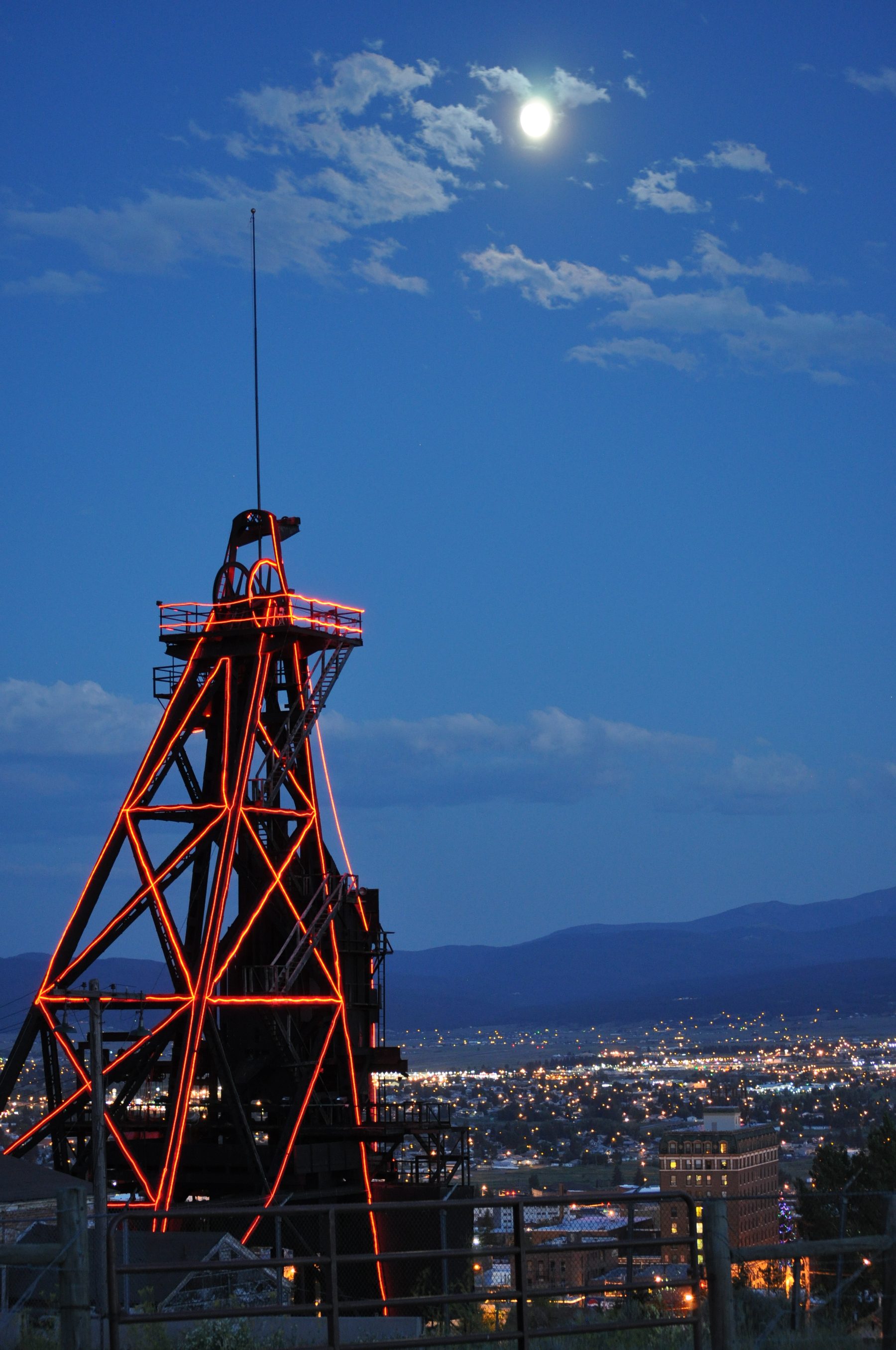 A headframe outlined in red light above the Butte valley at night, under a full moon